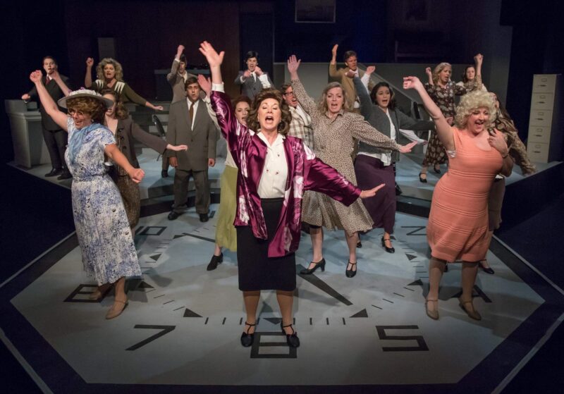 A scene with many women from the 9-to-5 Musical with different dresses and wigs with their arms up, singing